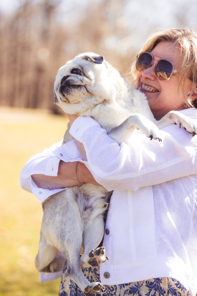DJ, a smiling woman with blonde hair and aviator sunglasses, joyfully hugs a cream-colored pug named Smooshie. She wears a white blazer and blue floral skirt. The sun-drenched outdoor setting features a blurred grassy field and bare trees in the background.