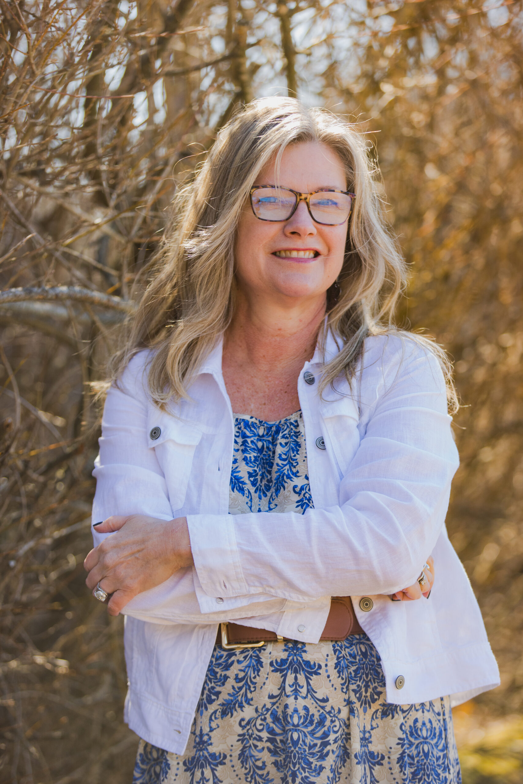 DJ, a smiling woman with blonde hair and tortoiseshell glasses, stands with arms crossed wearing a white blazer over a blue floral dress with a brown belt. She radiates warmth and confidence. Sunlit bare branches create a soft golden background behind her.