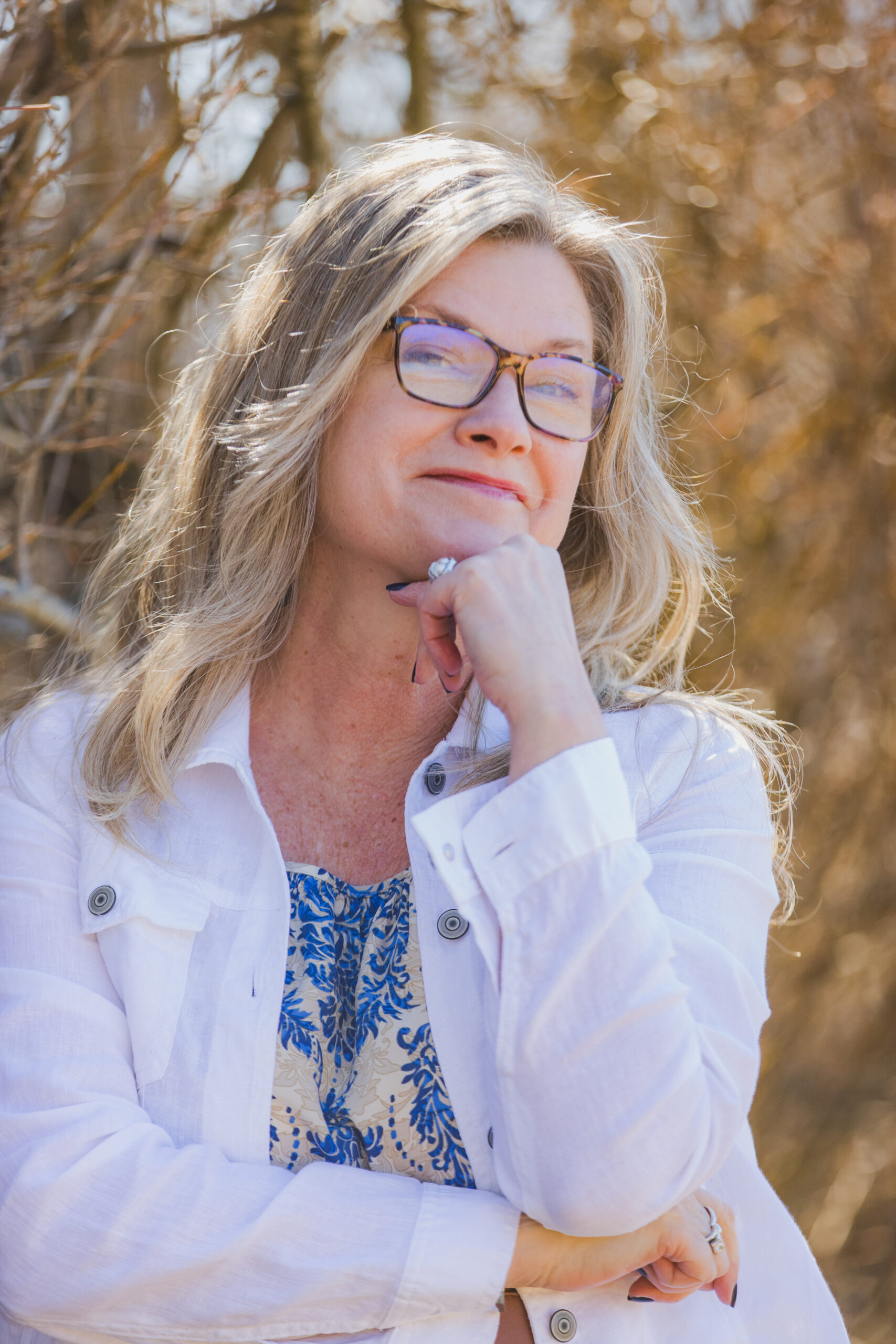 DJ, a woman with blonde hair and tortoiseshell glasses, gazes thoughtfully upward with her chin resting on her hand. She wears a white blazer over a blue floral top. Warm, sunlit bare branches are softly blurred in the background.