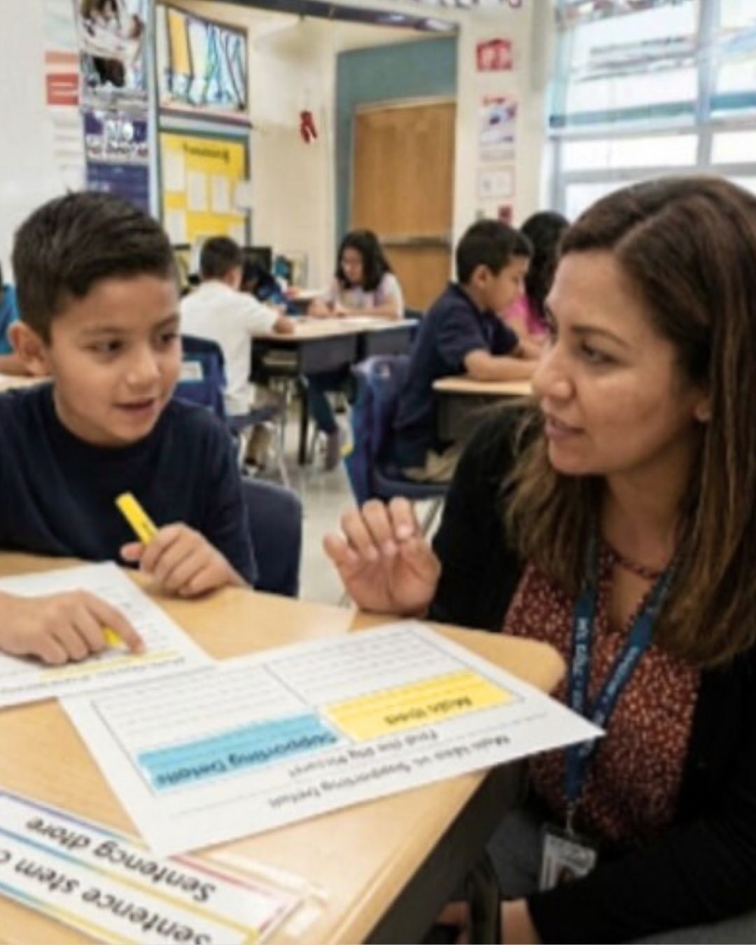 A teacher wearing a blue lanyard leans in to speak with a young student holding a yellow marker. Worksheets lay on the desk. Other students work at desks in the background of a decorated elementary classroom.