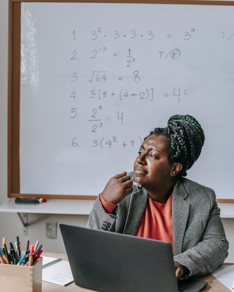 A teacher sits at a desk with a laptop, gazing upward in thought while holding a marker to her chin. Behind her, a whiteboard displays six math problems involving exponents and order of operations. Colored markers and a notebook are visible on the desk.
