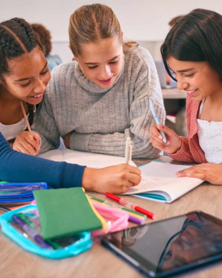 Three smiling middle school students huddle together over an open notebook at a desk, engaged in collaborative work. One student writes while the others look on. Colorful pencils, notebooks, a pencil case, and a tablet are spread across the desk. Other students are visible in the background.